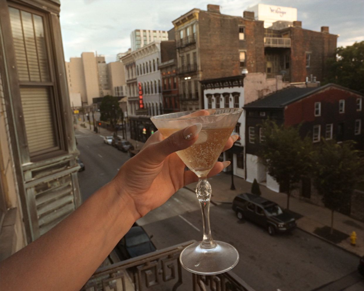 Person holding a cocktail on a balcony overlooking a city street. The drink sparkles in the sunlight.