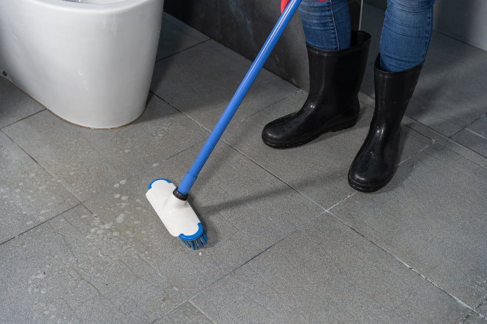 Person in rubber boots scrubbing bathroom floor with brush.