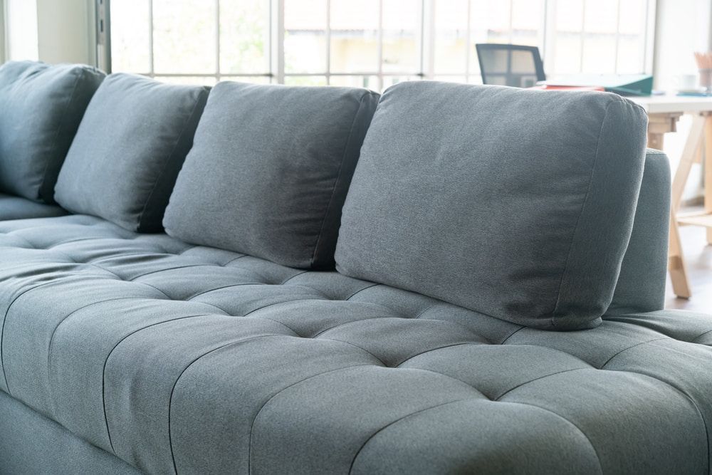 Gray tufted sectional sofa with three square pillows against a window.