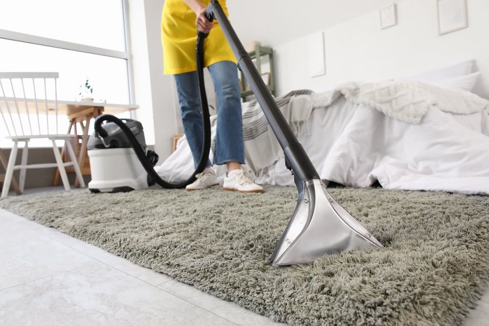 Person vacuuming a gray shaggy rug in a bright room with a white bed and chair.