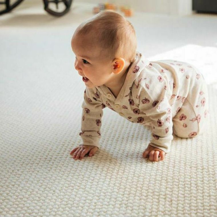 Baby in a patterned onesie crawling on a light carpet, looking to the side.