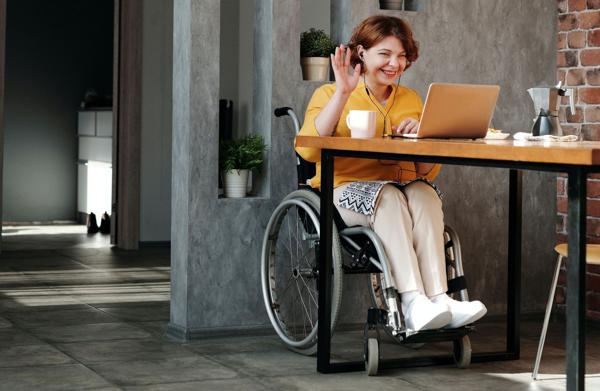 Woman in wheelchair waves at laptop during a video call at a table.
