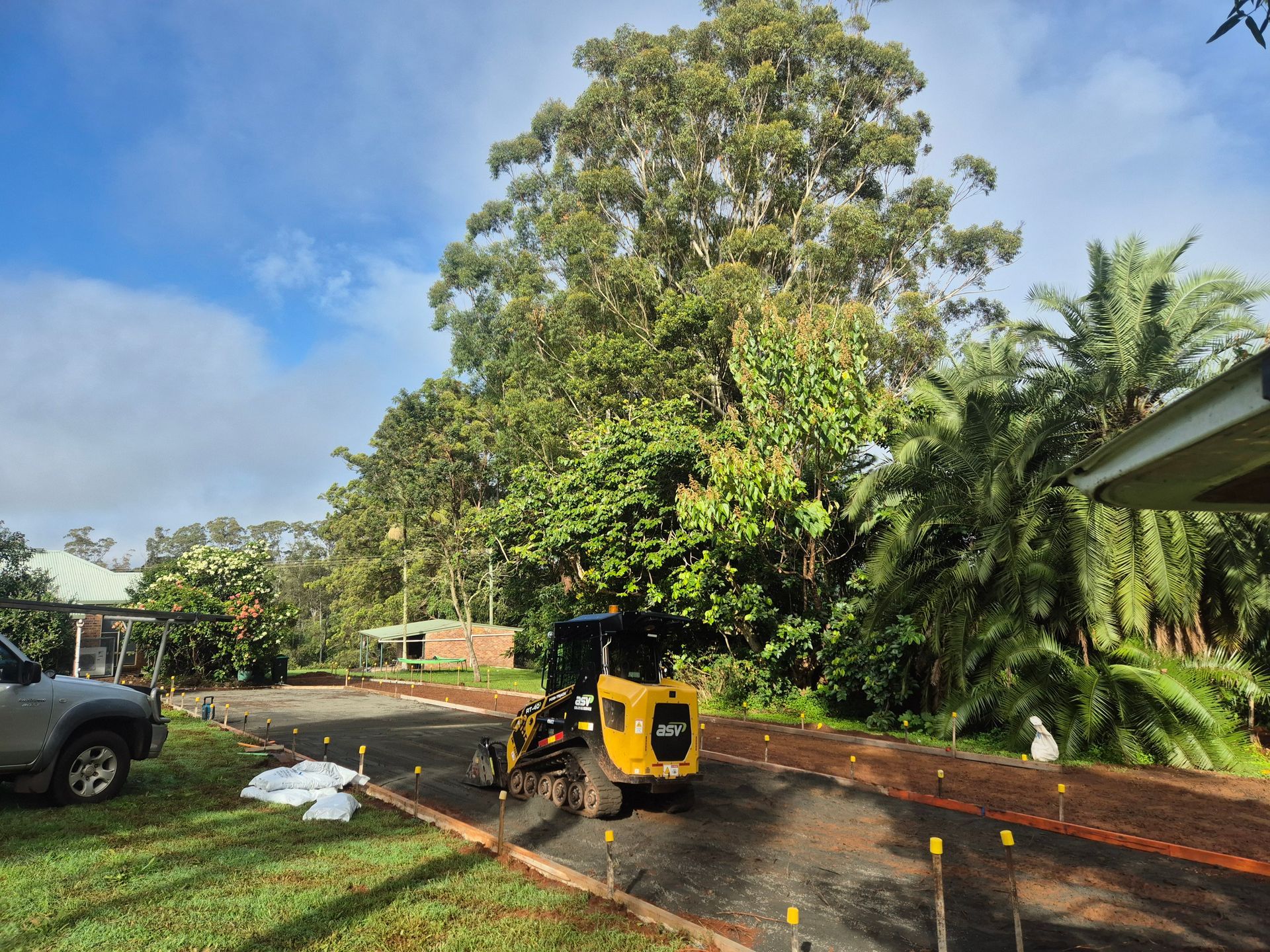 A Yellow Excavator on a Gravel Driveway, Preparing for Work Near a House and Trees — All Around Concreting in Wollongbar, NSW