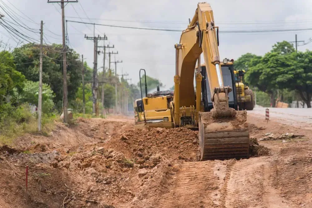 Yellow Excavator Digging a Trench on a Dirt Road, Next to Power Lines and Trees — All Around Concreting in Ballina, NSW