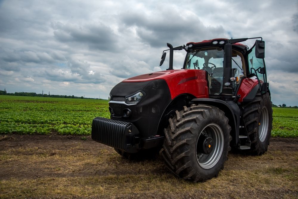 Red tractor parked in a muddy field under a cloudy sky, with green crops in the background.