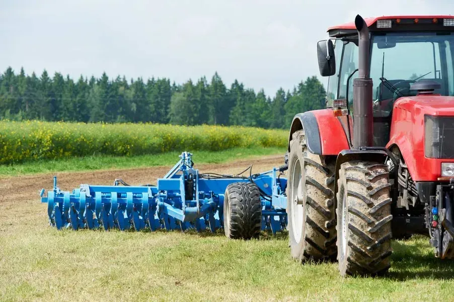 Red tractor pulling a blue farm implement across a grassy field beside trees and crops. — Bundaberg Field Service in Bundaberg East, QLD
