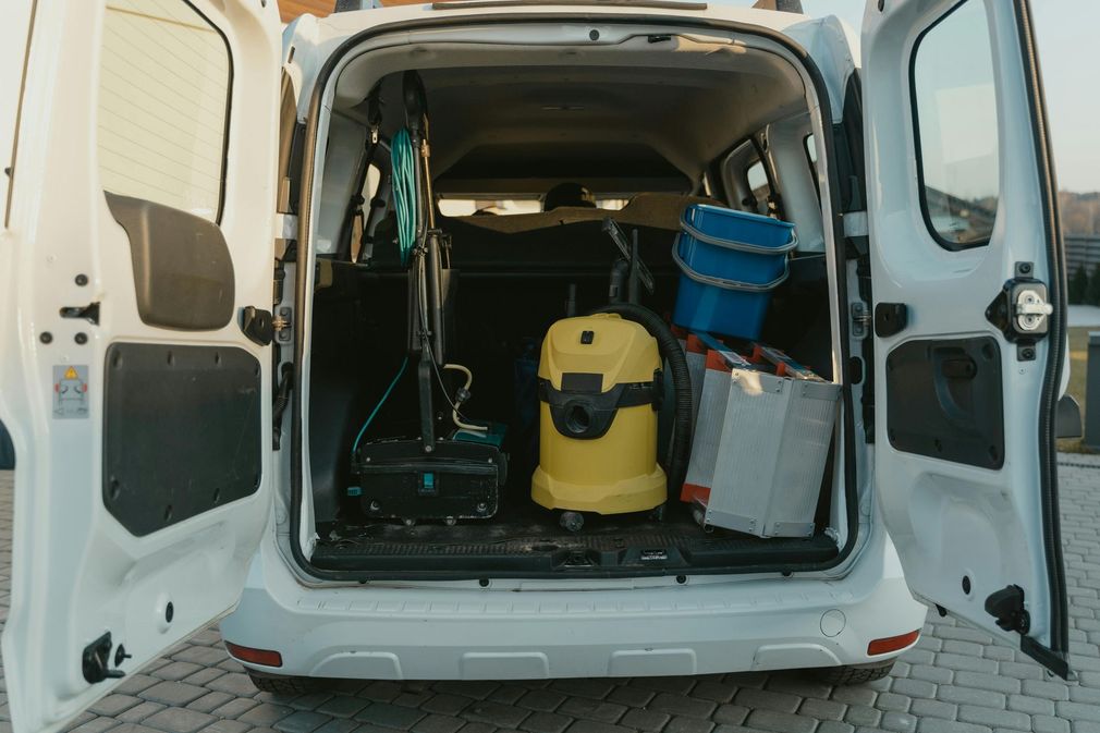 White van open, revealing cleaning supplies: vacuum, buckets, tools. Outdoors, daytime.