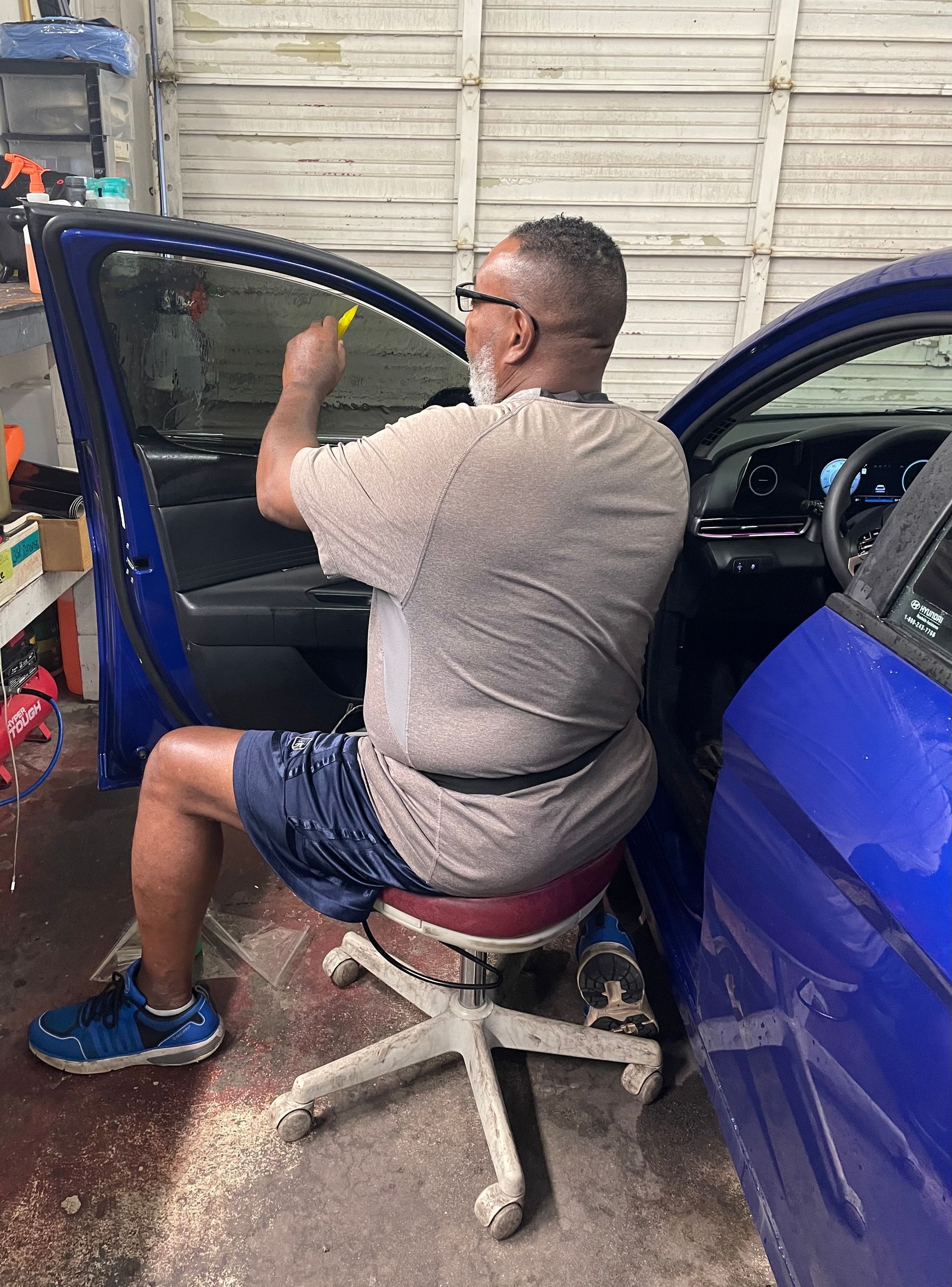 A man is sitting on a stool in front of a blue car.