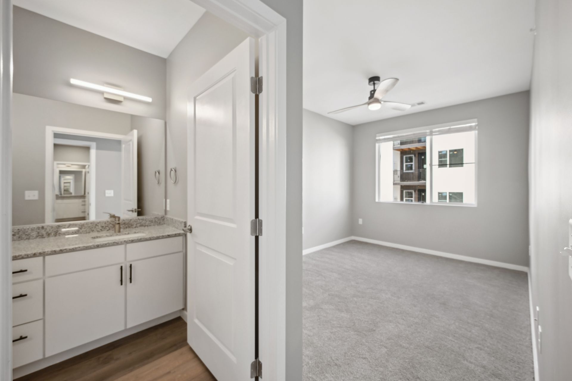 A view of a modern bathroom with grey vanity and white door, opening into a bright bedroom with light grey carpet.