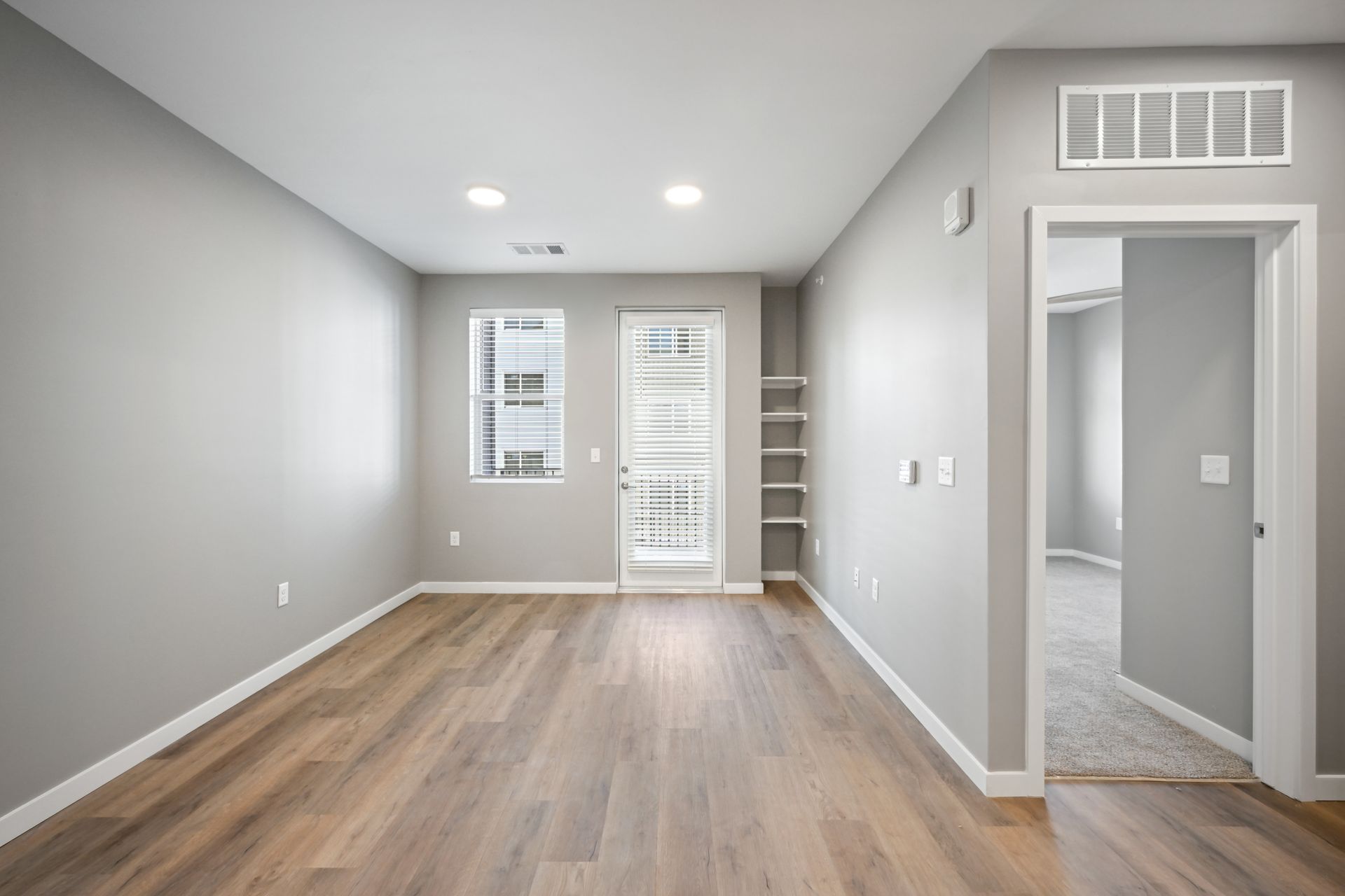 A brightly lit, unfurnished room with gray walls, light wood-look flooring, a glass door, a window, and a doorway.