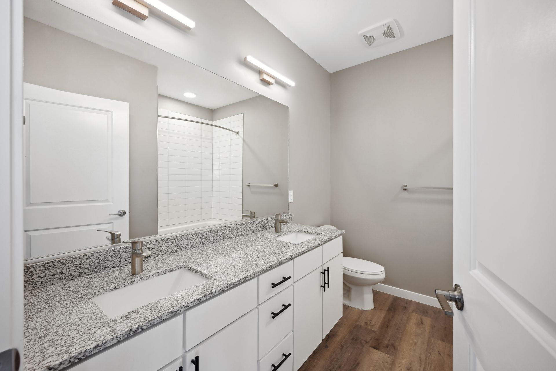 A modern bathroom with a granite vanity, white cabinets, a large mirror, a toilet, and wood-look flooring.