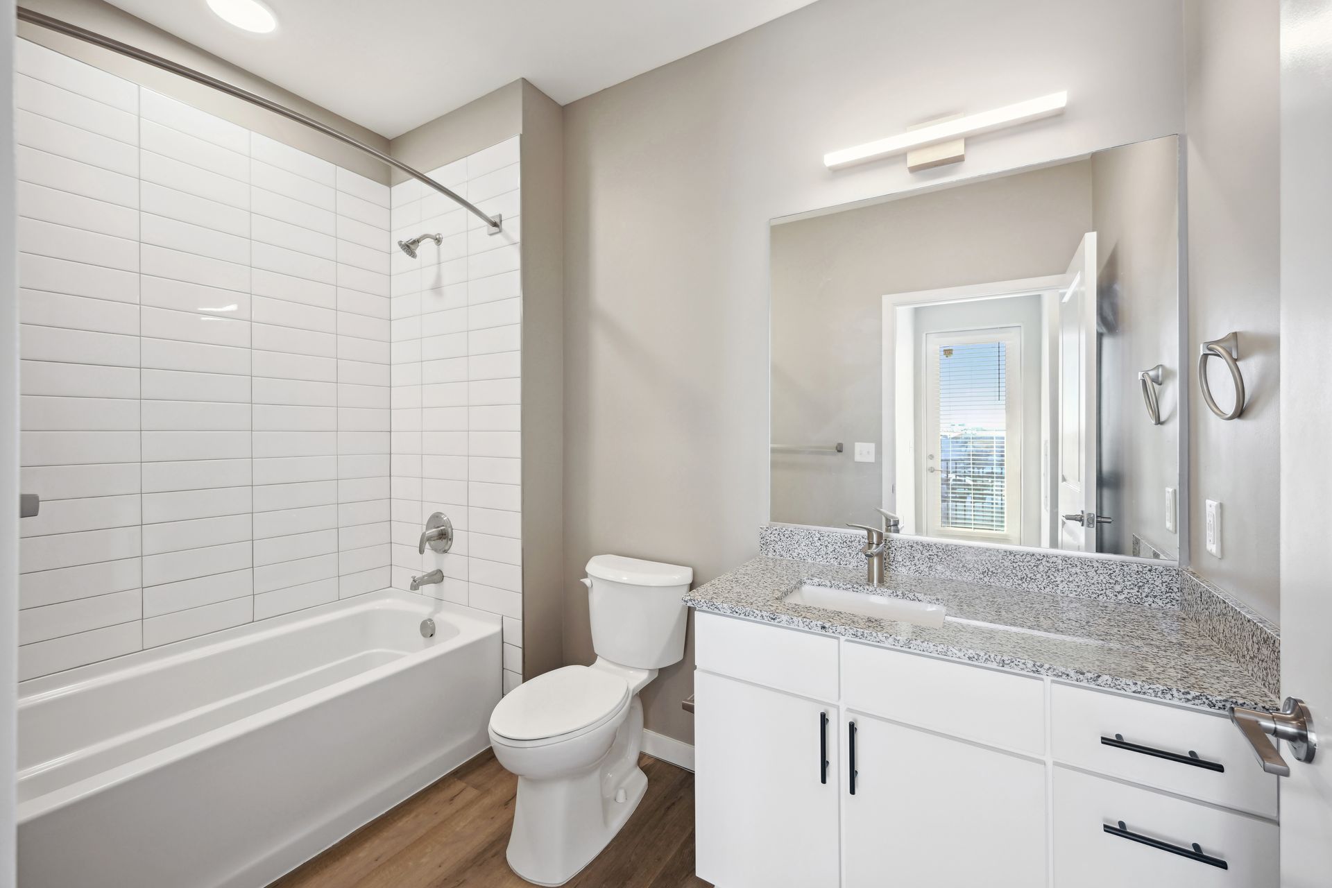 Modern bathroom featuring a white tiled bathtub, a toilet, a granite vanity with a white cabinet, and a large mirror.