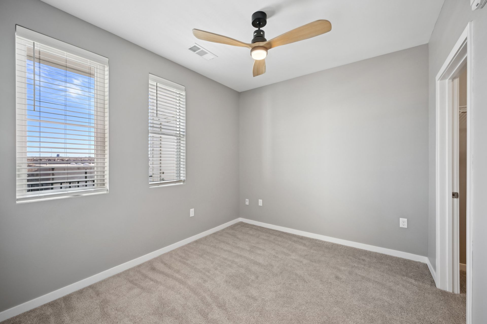 An empty bedroom with gray walls, beige carpet, two windows with blinds, and a ceiling fan with light wood blades.