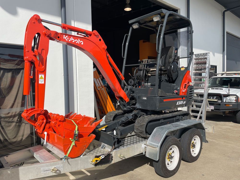 A Small Red Excavator is Parked on a Trailer in Front of a Building — Adzcom Civils in Portsmith, QLD