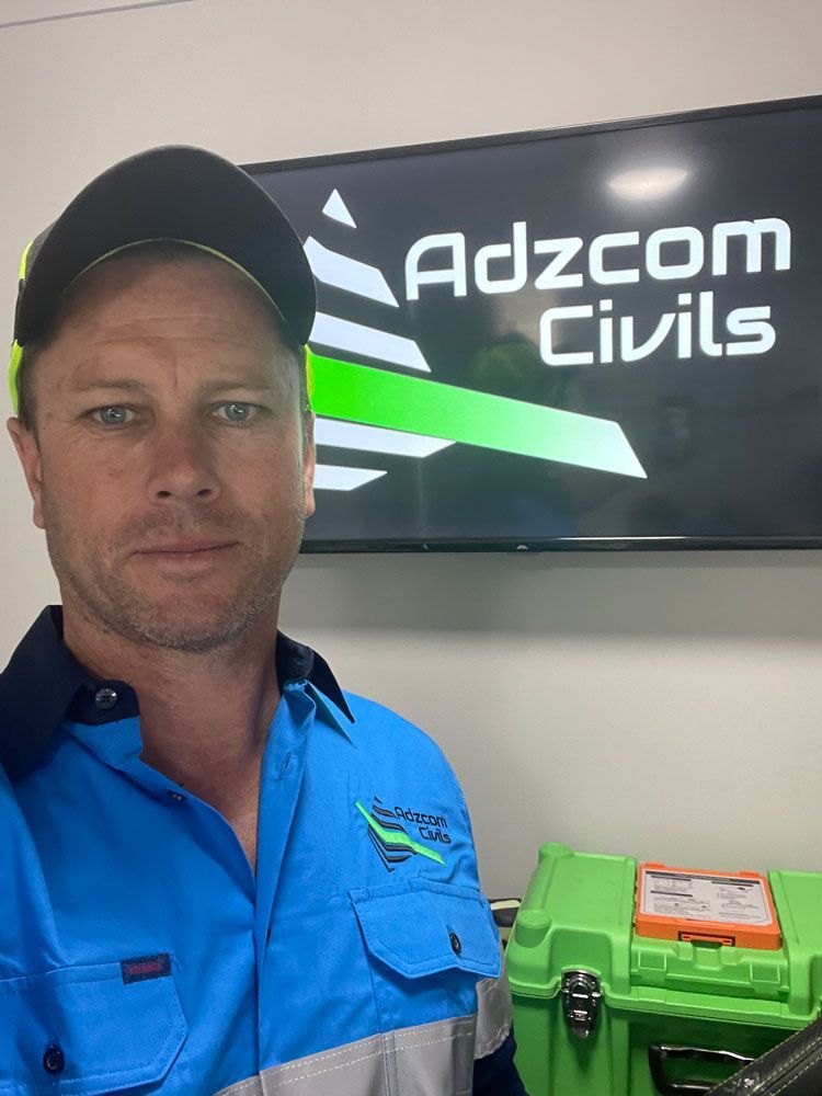 A Man in a Blue Shirt is Standing in Front of an Adzcom Civils Sign — Adzcom Civils in Innisfail, QLD