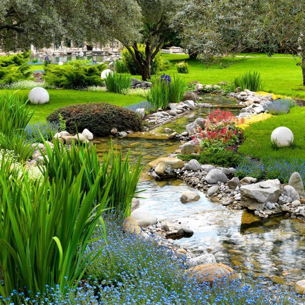 A peaceful garden with a flowing stream, lush green grass, rocks, and colorful flowers.