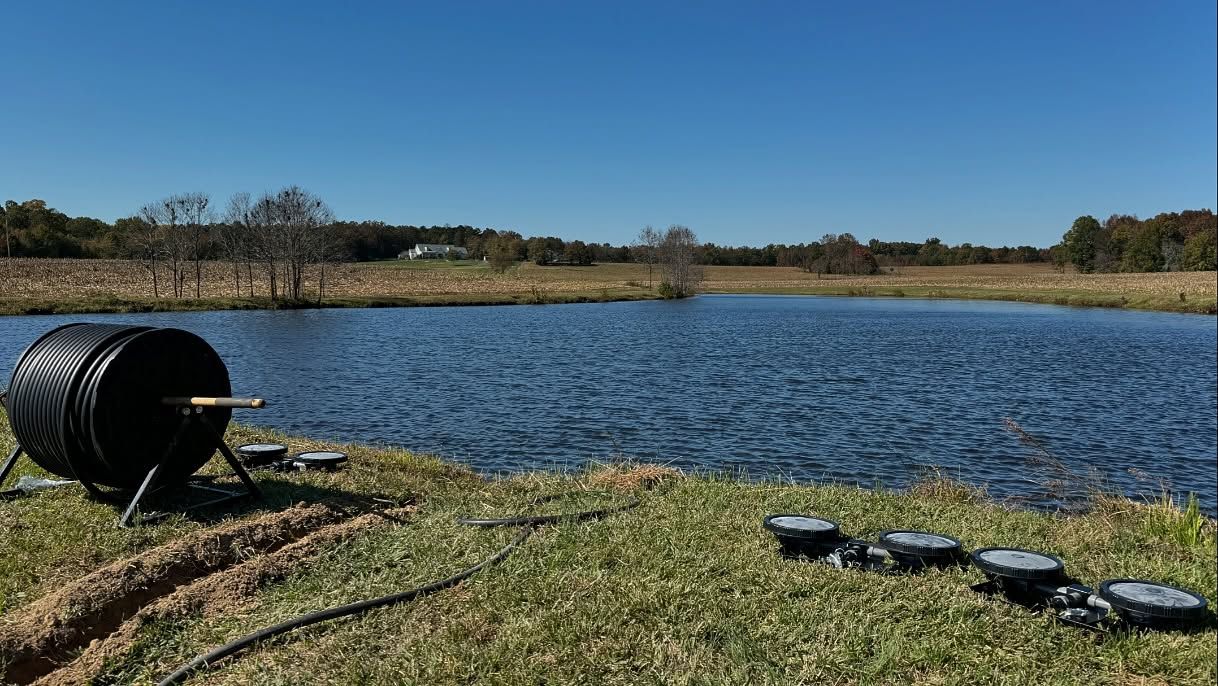 A black pond aerator with tubing sits by a calm pond in a grassy field.