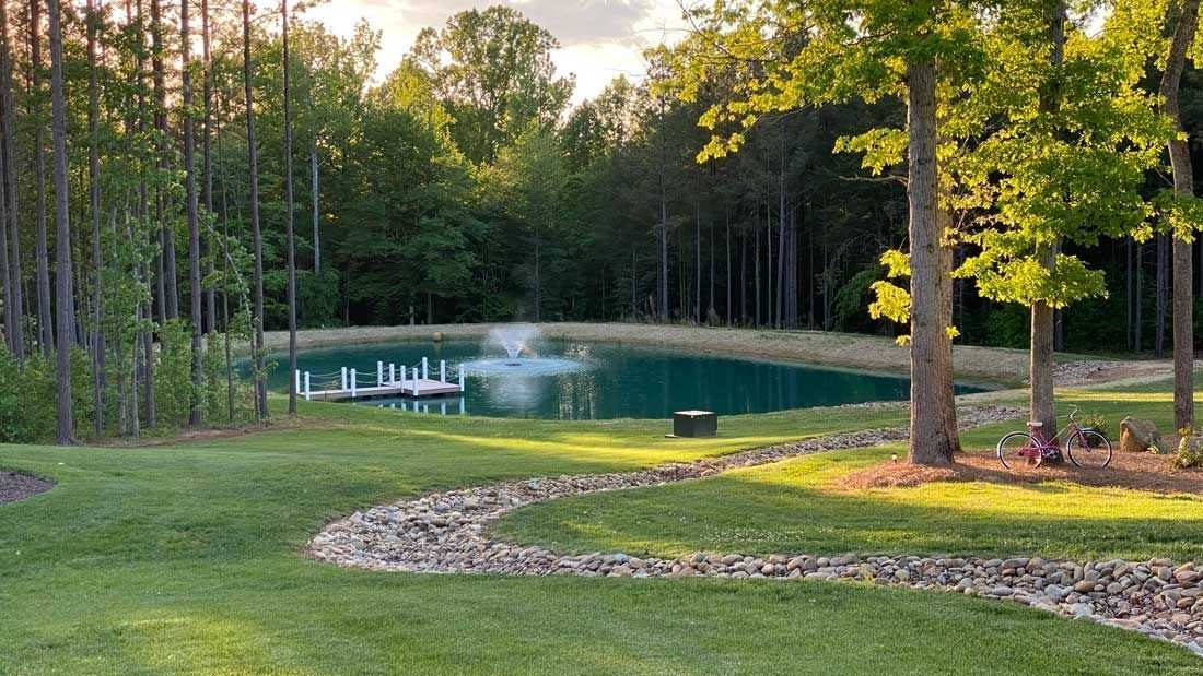 Lush green yard with a winding rock path leads to a pond with a fountain, surrounded by trees.