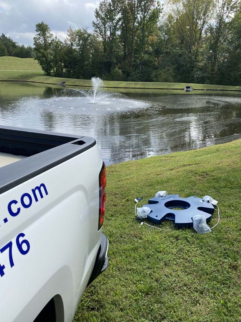 White truck beside a pond with a fountain, and a blue, flat fountain base on the grass.