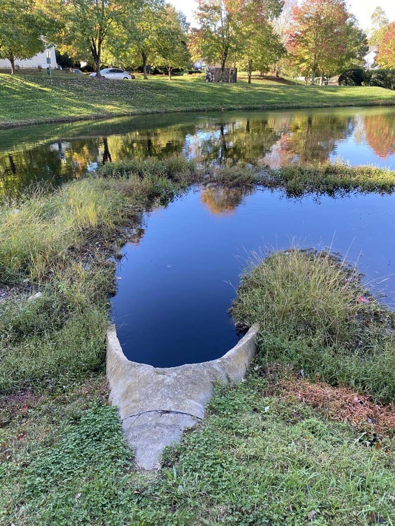 Concrete pipe draining into a pond with a grassy bank, trees with fall foliage, and reflections in the water.