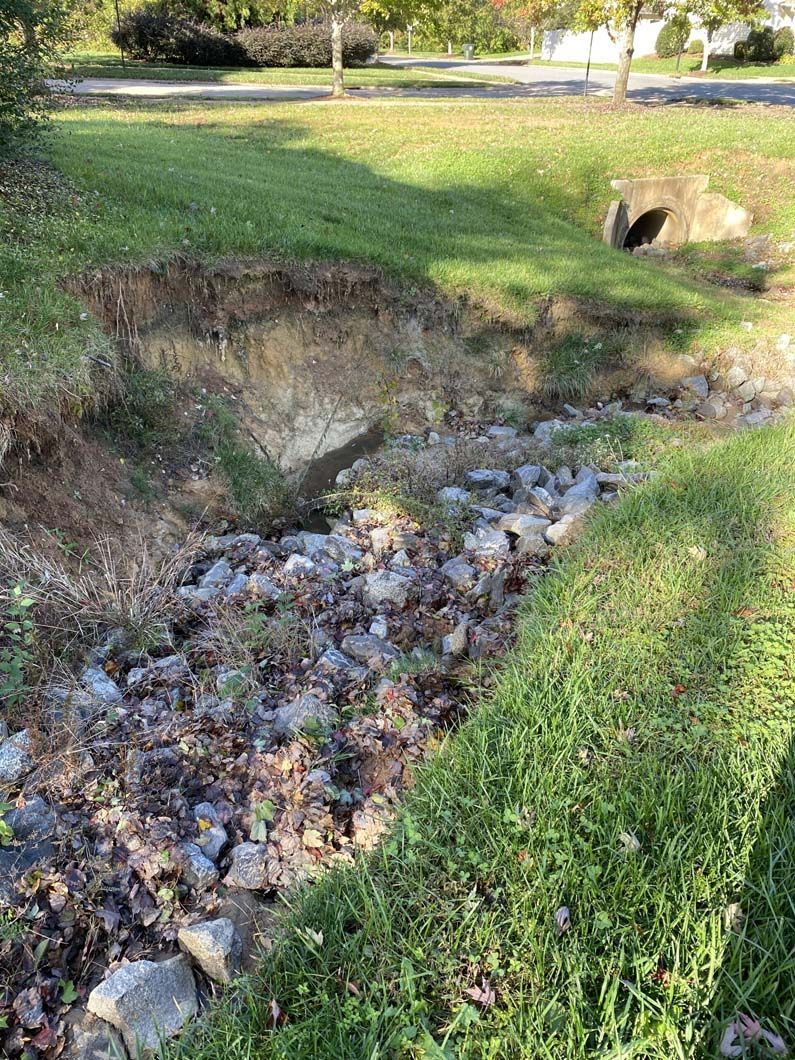Eroded bank of a small stream; grass and rocks line the edges leading to a culvert opening.