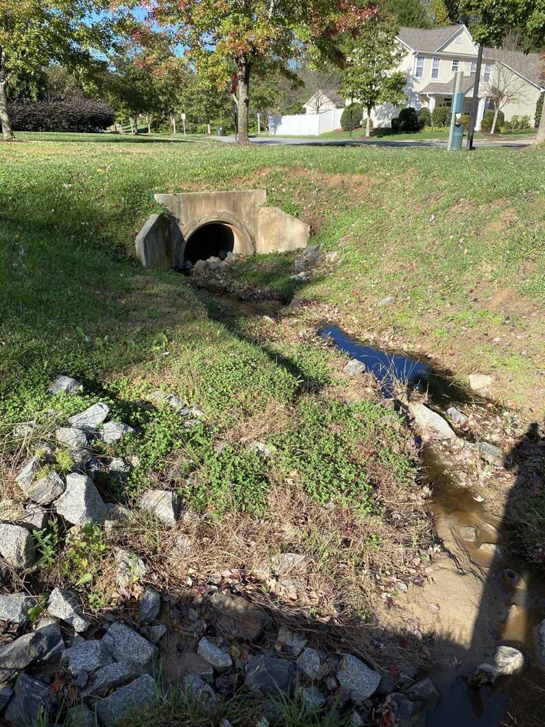 A concrete culvert tunnel entrance in grassy area. A small stream flows out from tunnel to the right. Sunny day.
