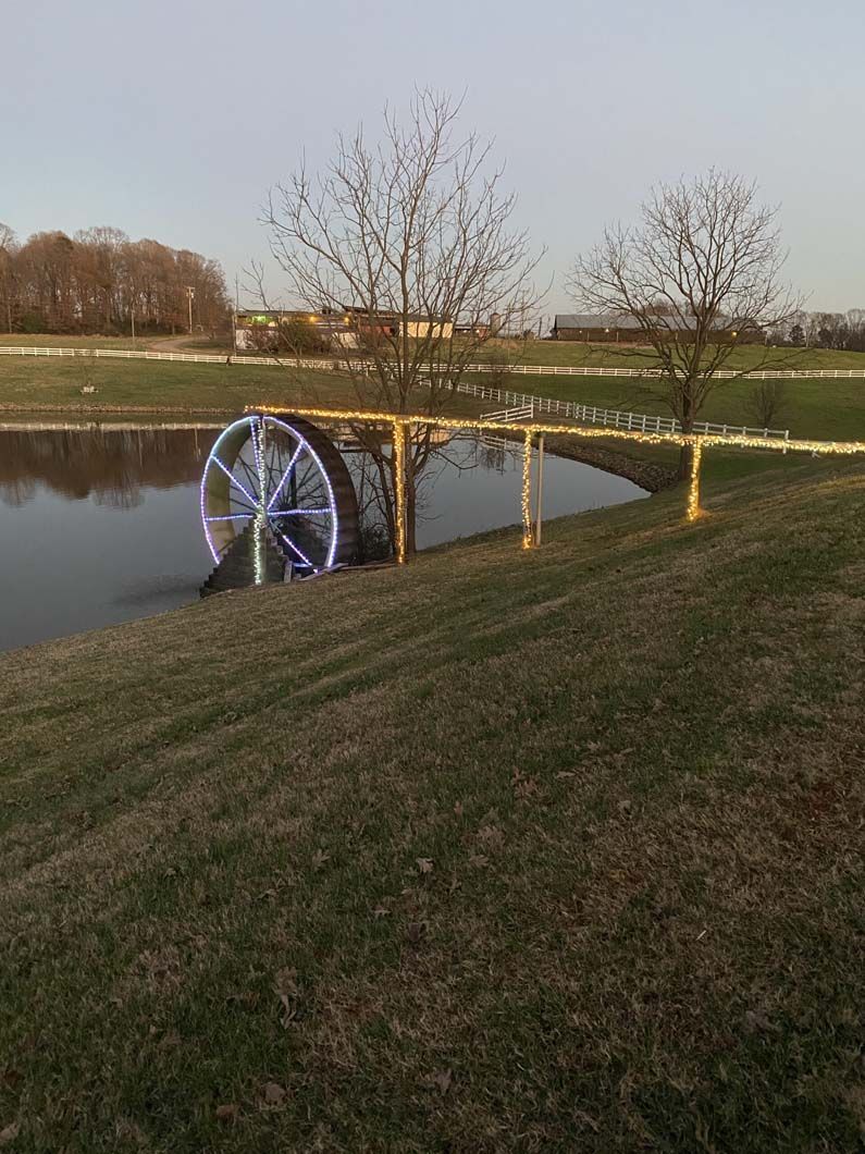 A glowing water wheel in a pond, with bare trees and grassy banks at dusk.