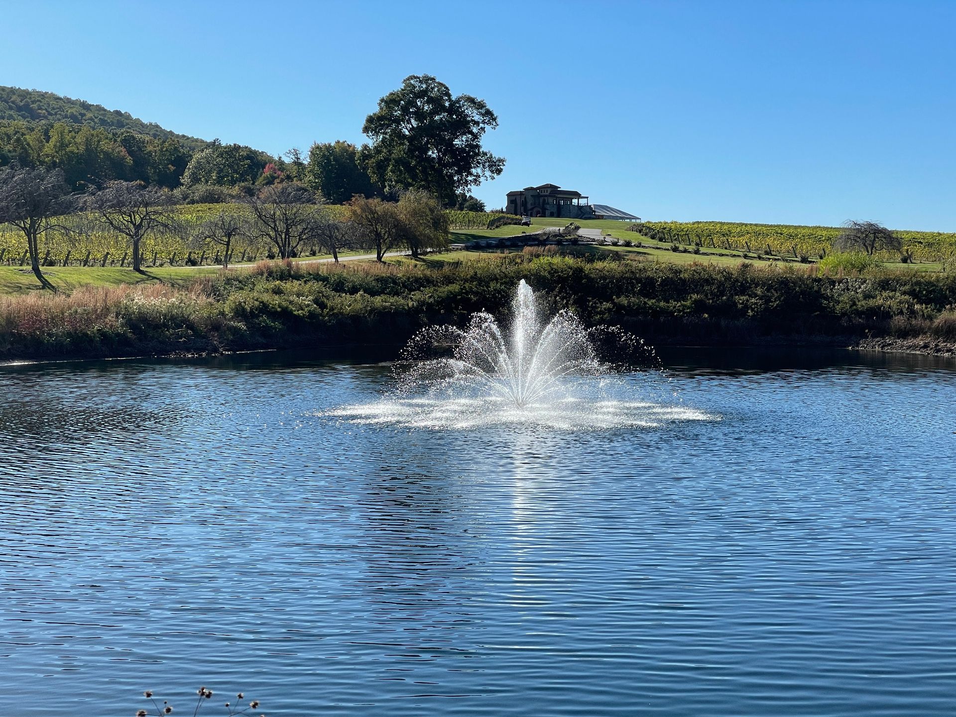 Fountain sprays water in a blue lake with a hillside and clear sky in the background.