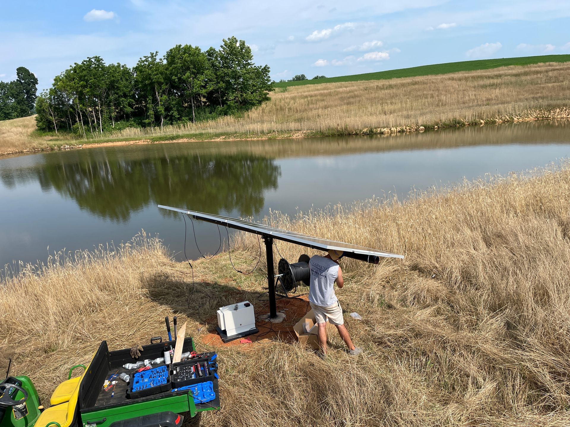 Man adjusting solar panel by a pond, with a tractor in the foreground.