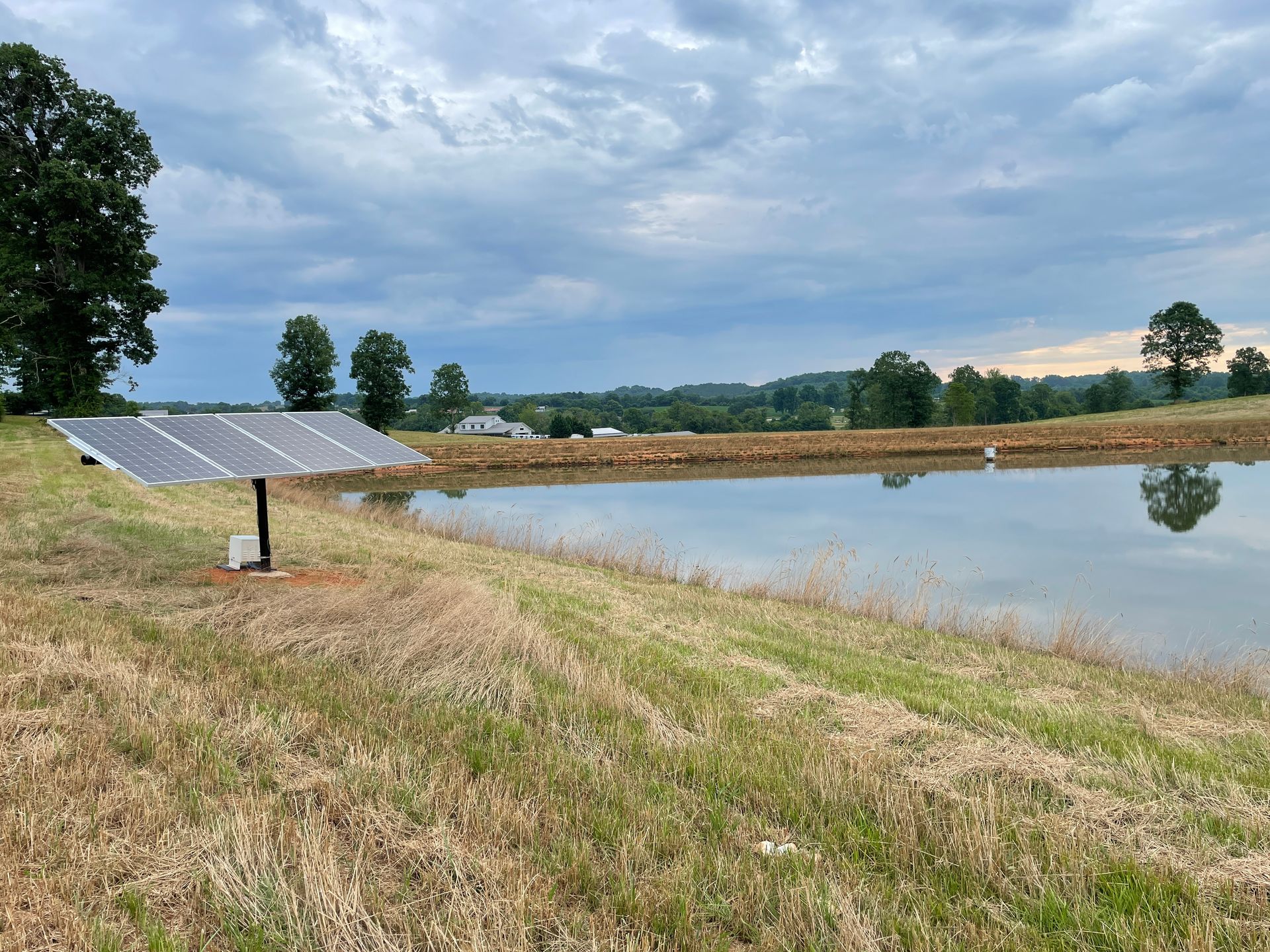 Solar panel over a pond in a field; trees and cloudy sky in the background.
