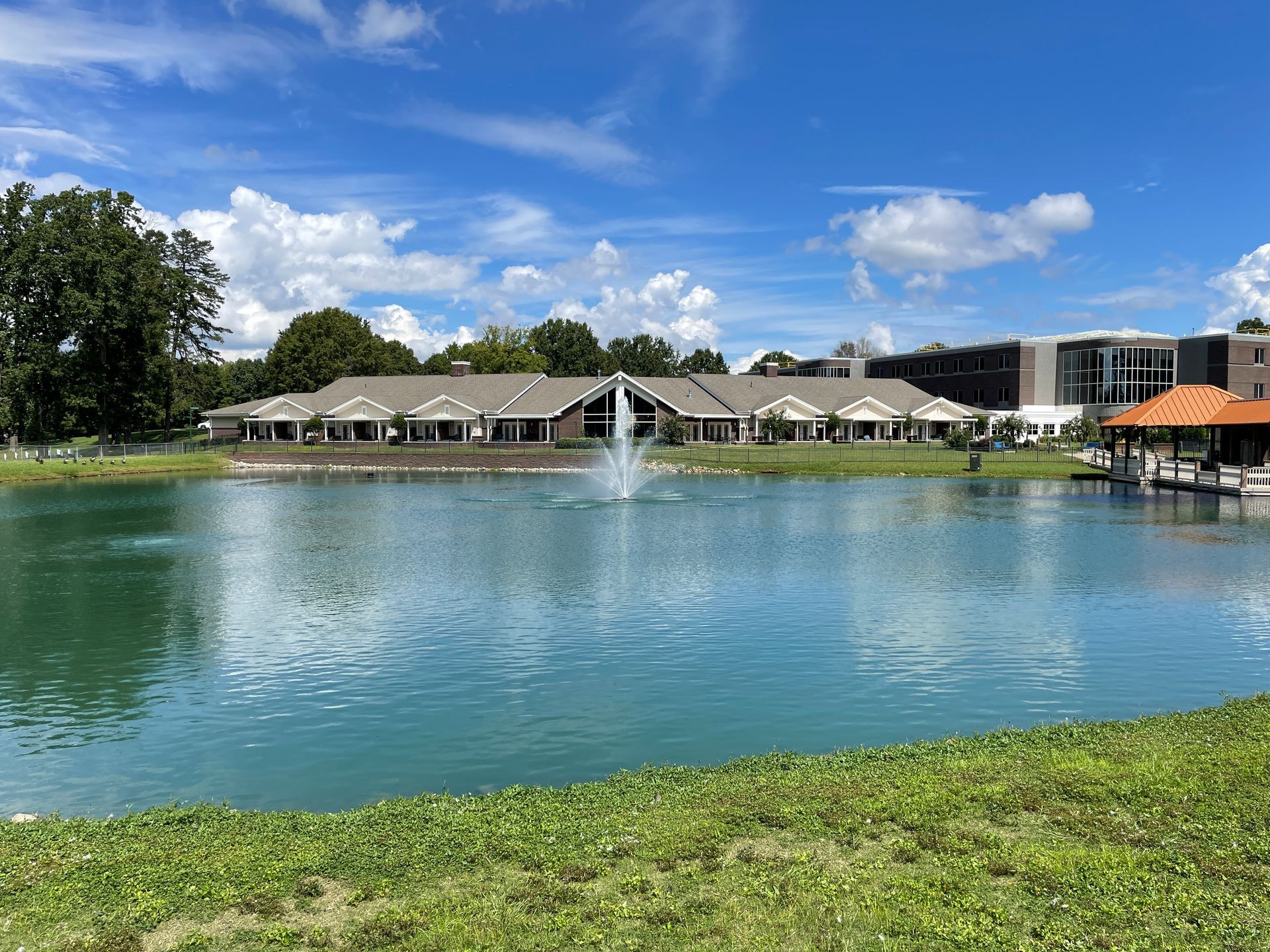 Pond with fountain in front of a building on a sunny day. Blue water, green grass, and sky.