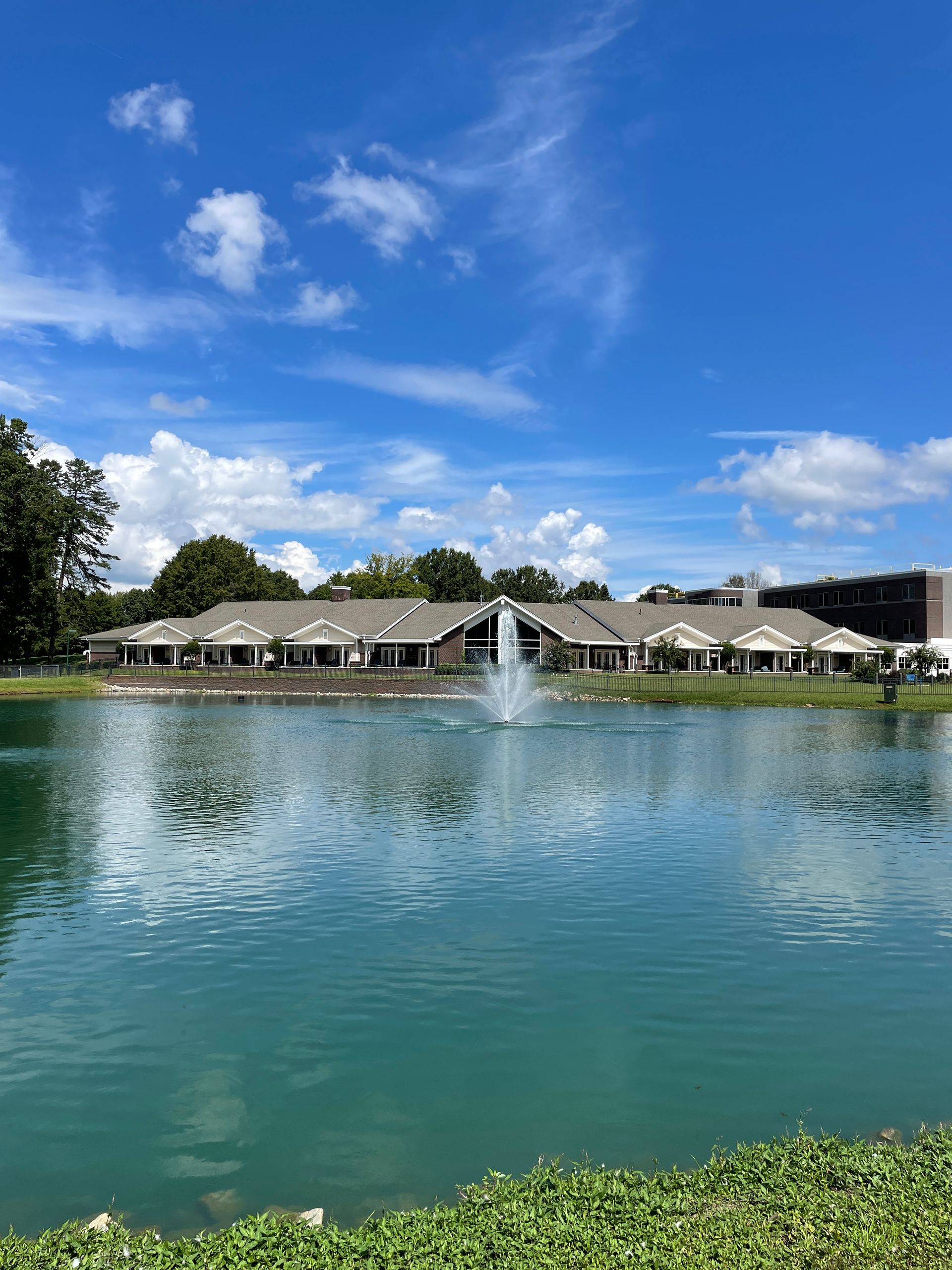 A teal pond with a fountain in front of a long building under a blue sky with clouds.
