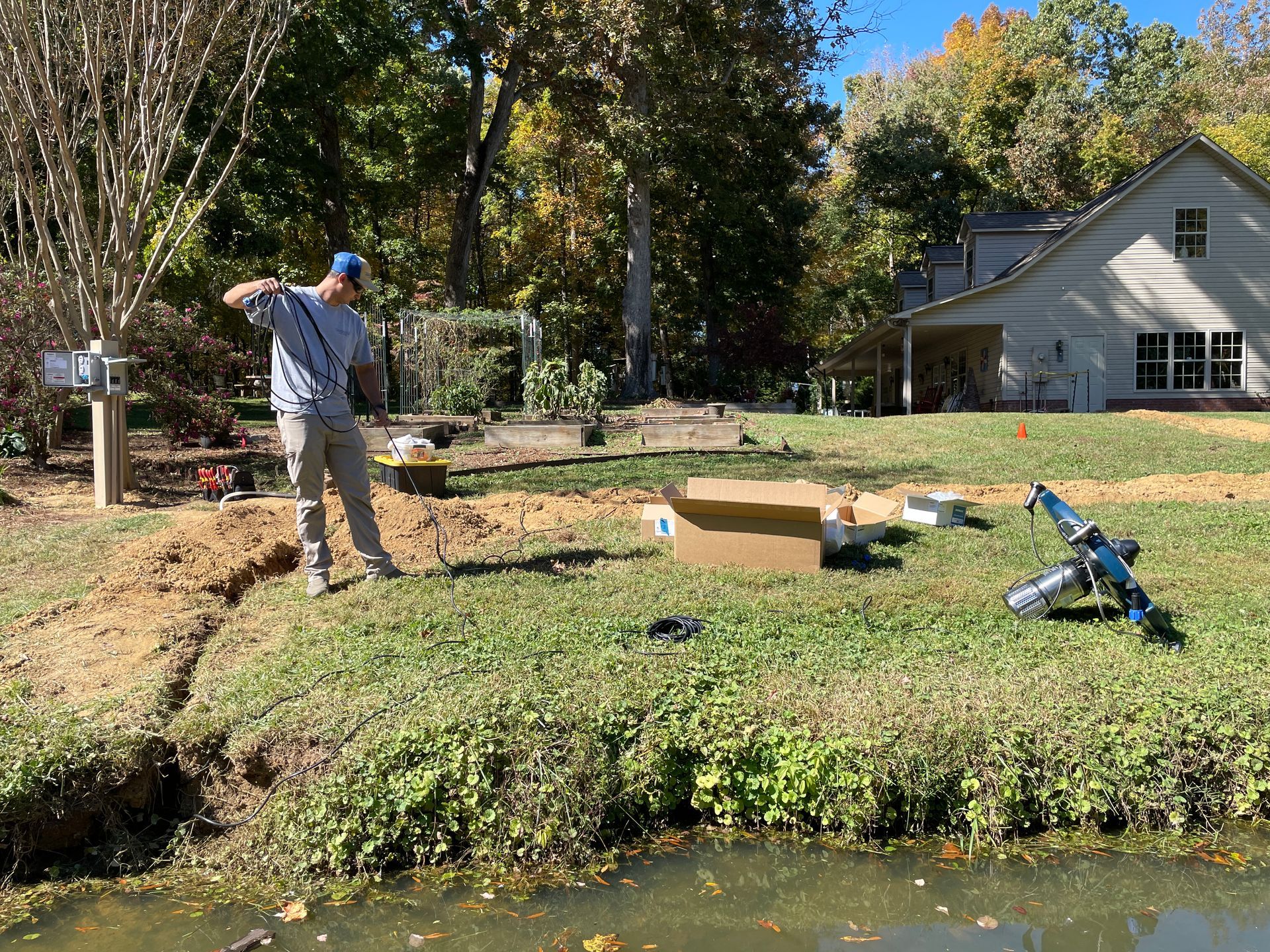 Man working outdoors near a waterway, digging a trench near a house, with tools and supplies nearby.