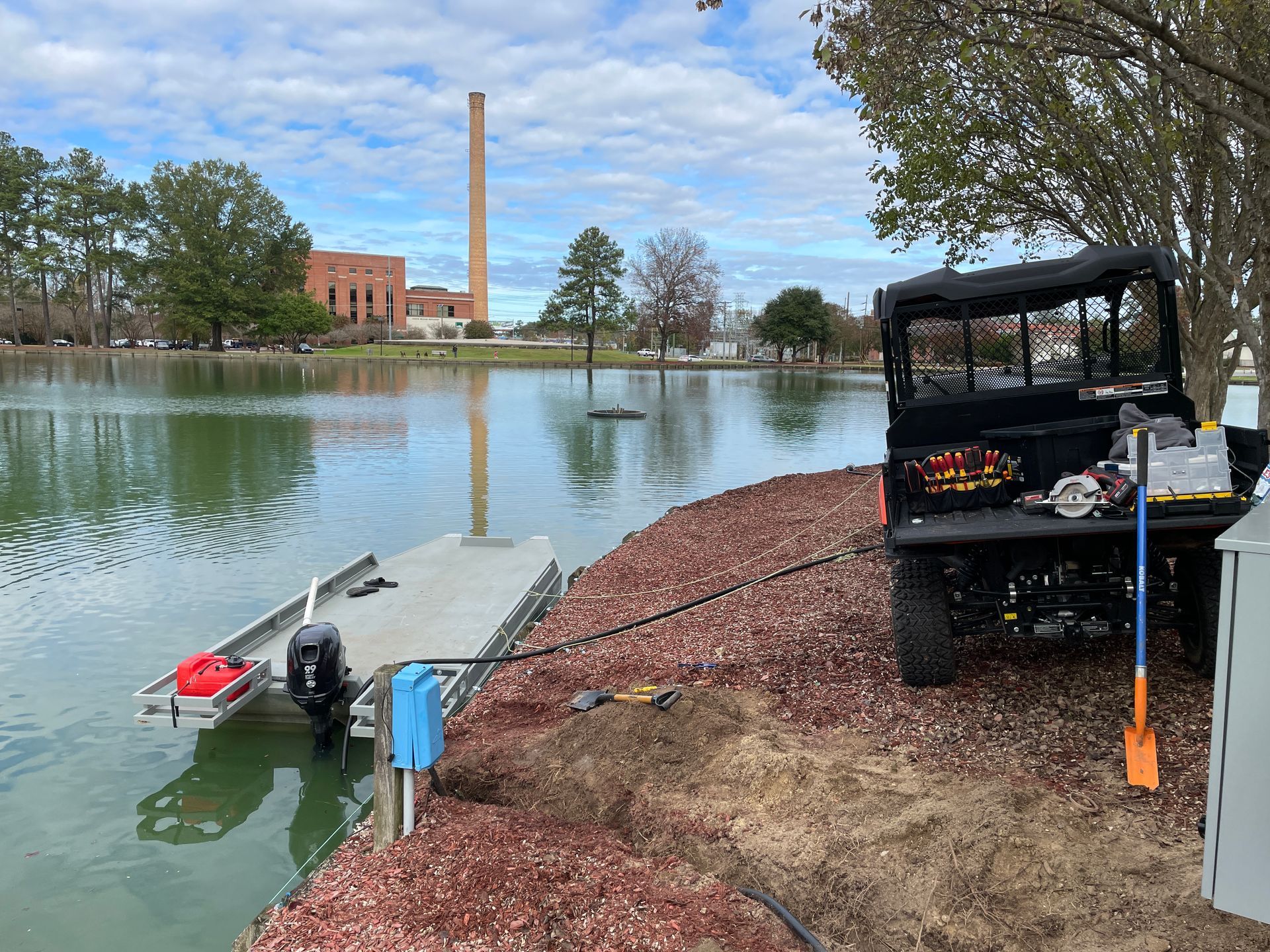 Utility vehicle near a pond with a dock. A tall smokestack and building in the background.