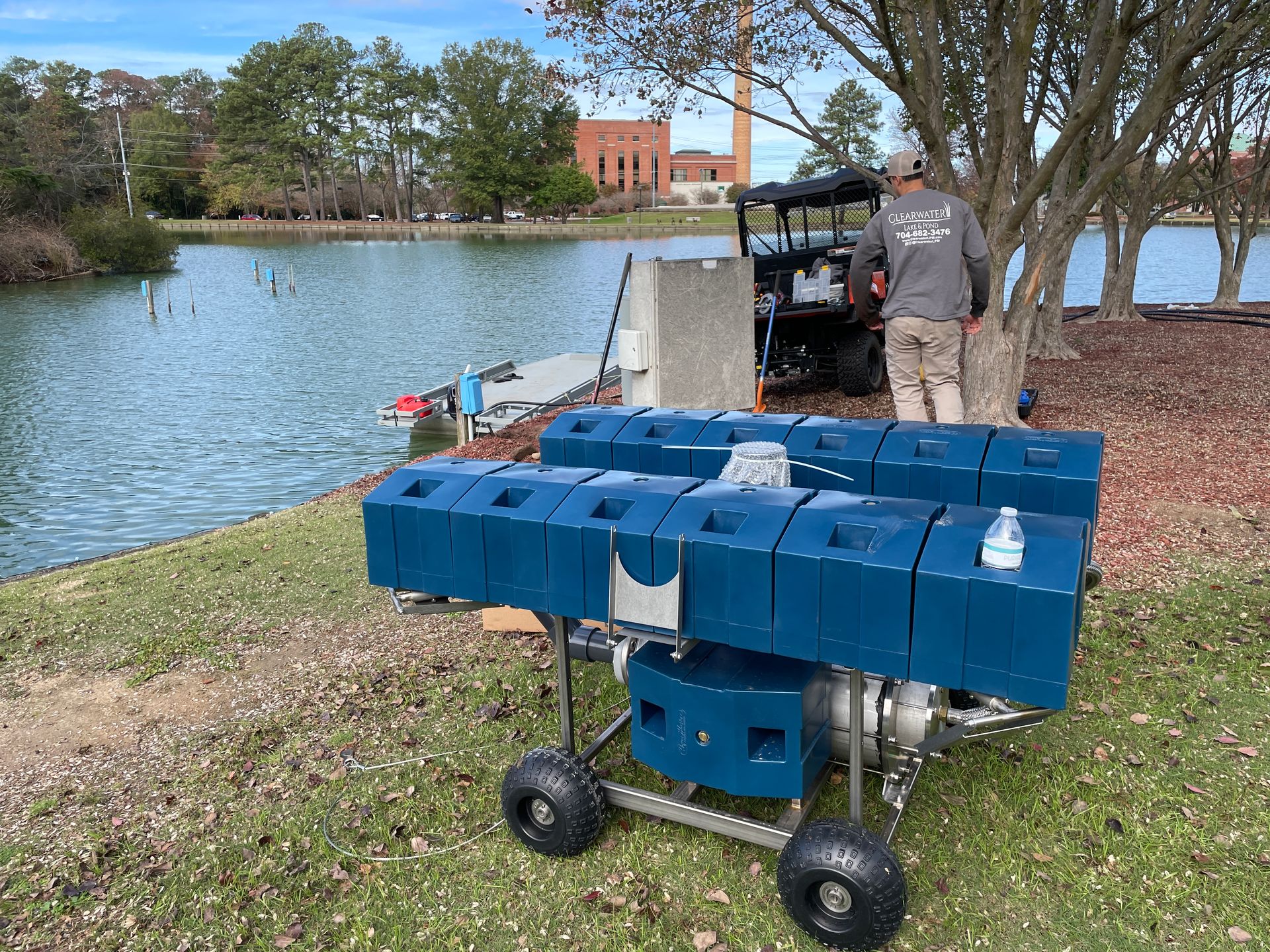 Man by lake with equipment. Blue machines on wheels sit on the bank; golf cart in background.