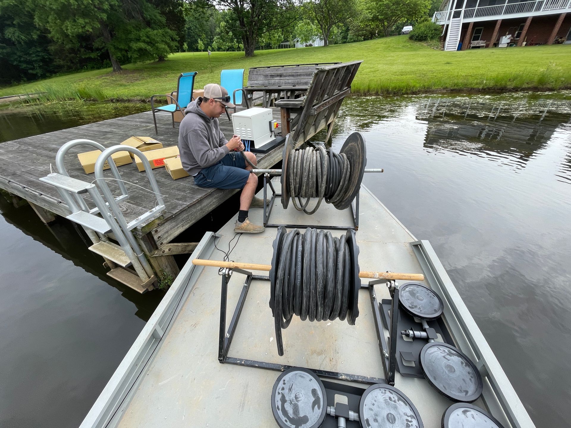 Man sits on dock by pond working on equipment with coiled hoses.