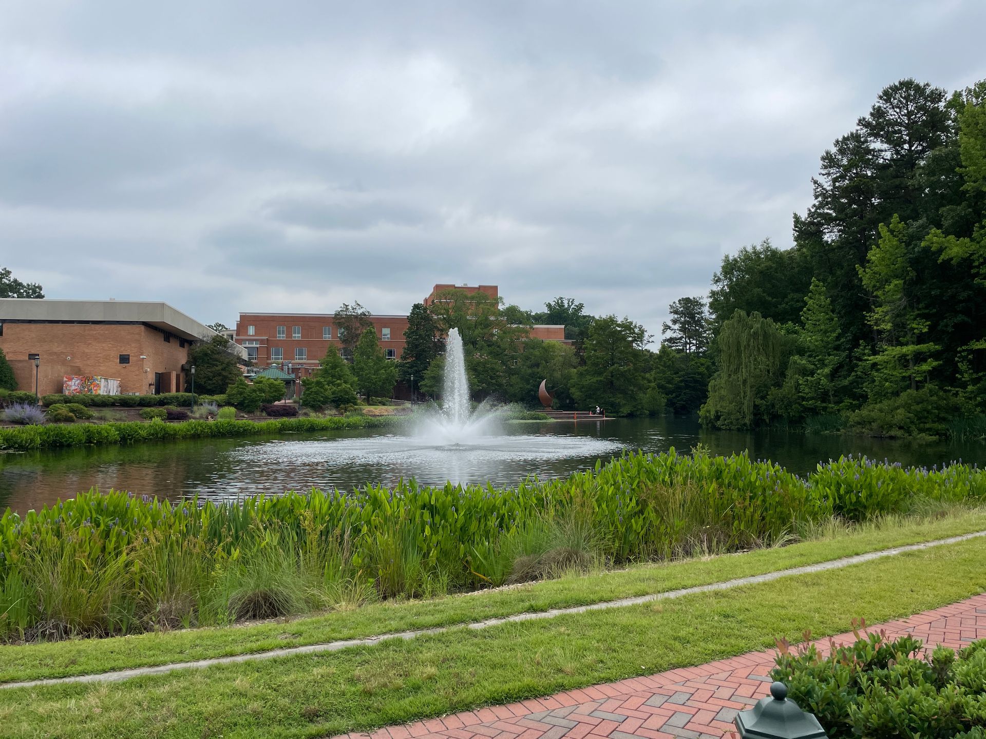 Fountain sprays water in a pond surrounded by greenery and brick buildings under a cloudy sky.