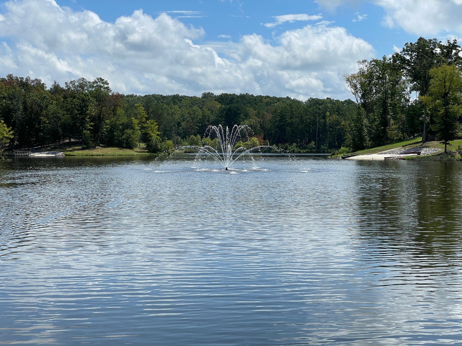 Lake with fountain in the center, surrounded by trees and a cloudy blue sky.