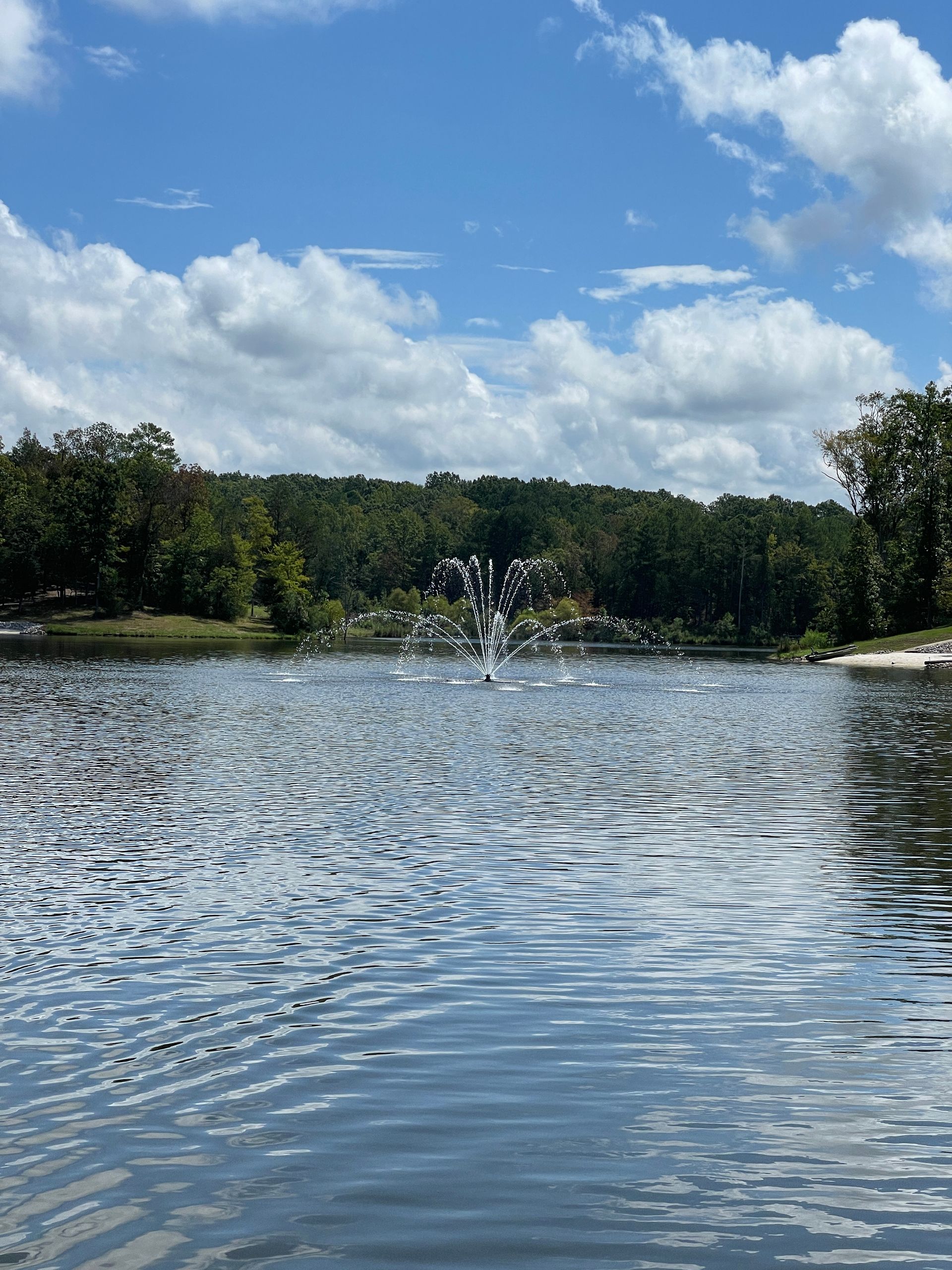 Lake scene with fountain spraying water; trees in the background, blue sky and puffy white clouds.