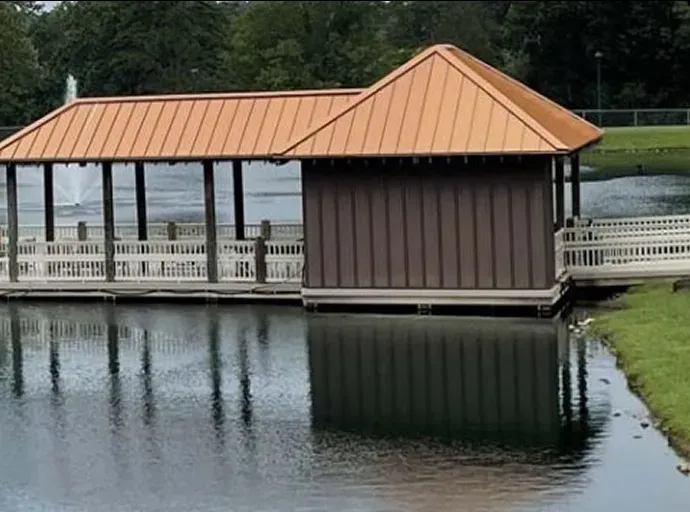 Brown boathouse on a lake with reflective water. Copper roof, wooden posts, and a white walkway.
