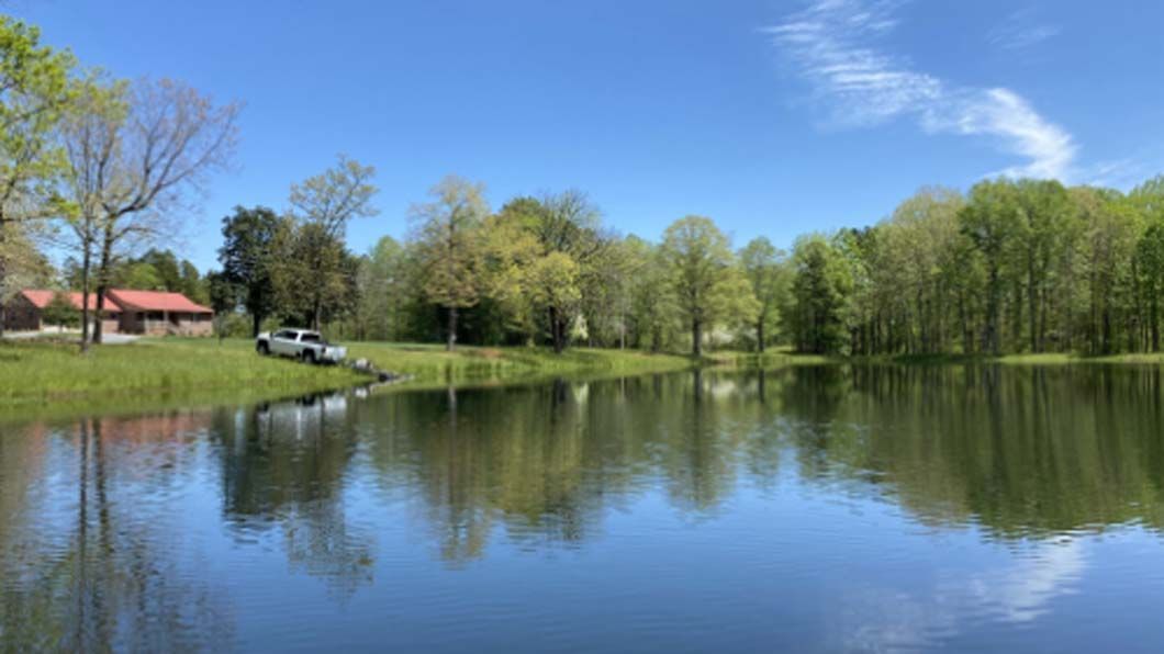 Calm lake reflects trees under a bright blue sky; a building and truck are visible on the shore.