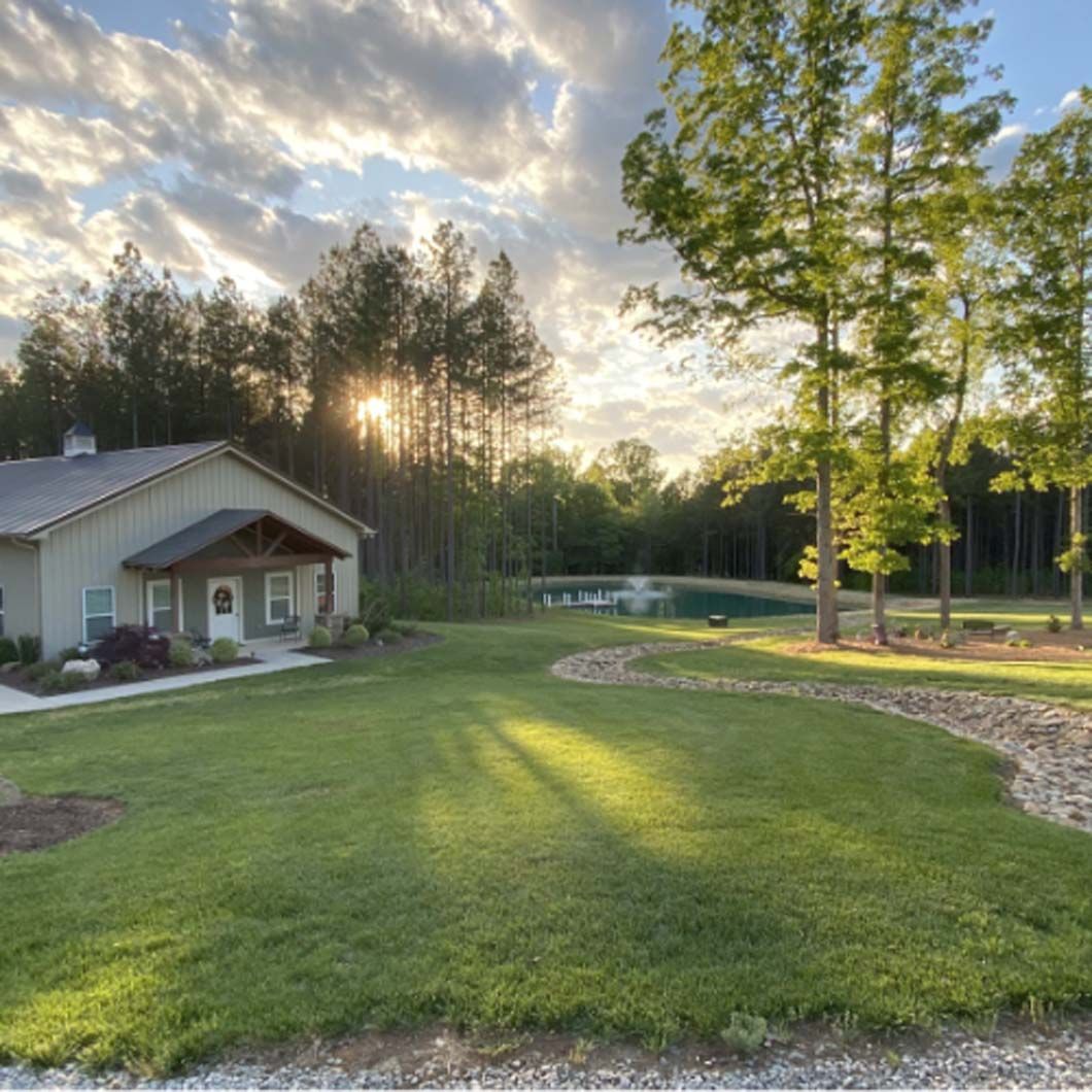 A building and lawn with trees, sunlight, and a pond in the background.