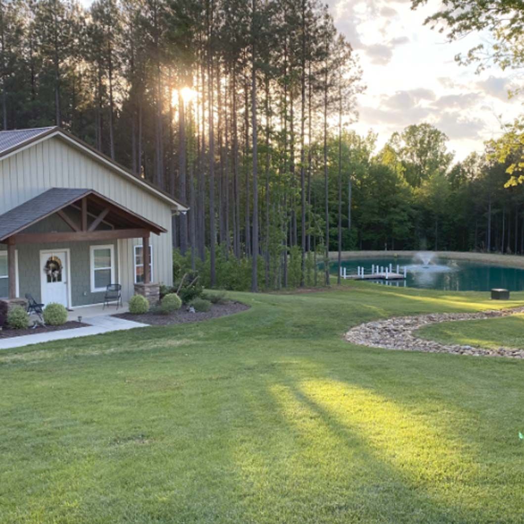 A house with a front porch, a pond, and tall trees; sunny outdoor scene.