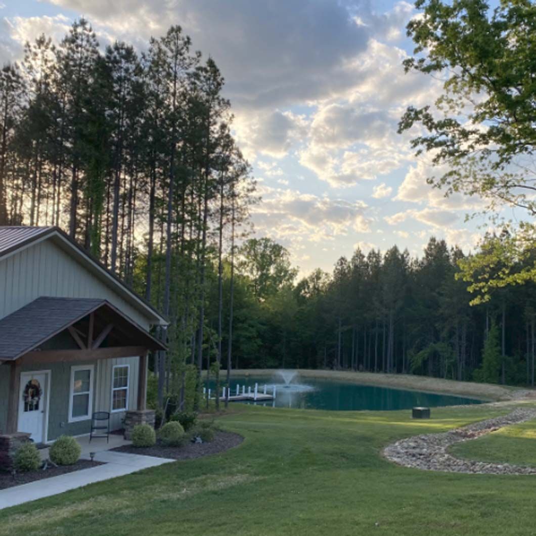A green house with a porch next to a pond with a fountain, trees, and a cloudy sky.