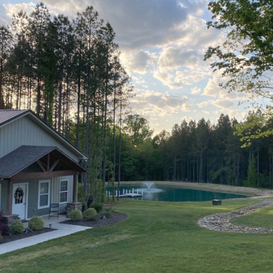 House with a pond in a wooded area. Green grass, blue water, and a cloudy sky.