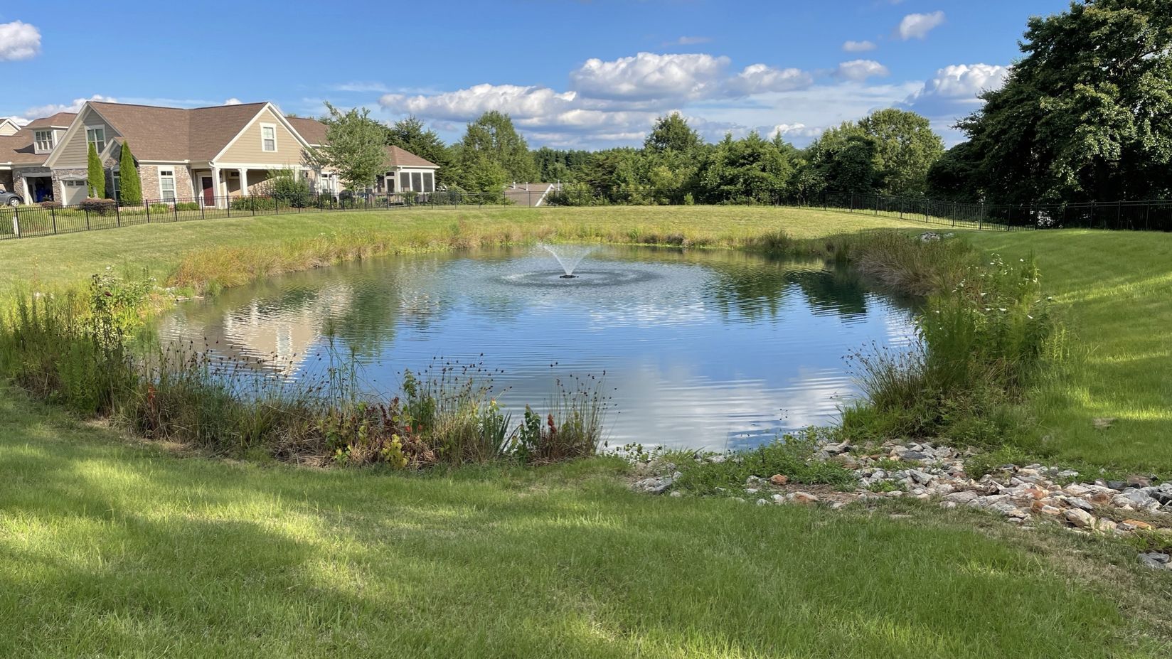 Pond with fountain and grassy banks, reflecting sky. Houses in background, sunny day.