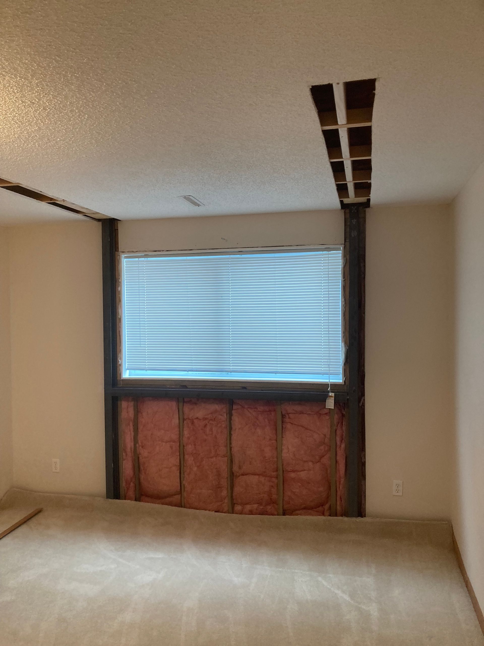 A room interior featuring a window with exposed insulation beneath it and partially opened drywall along the ceiling.