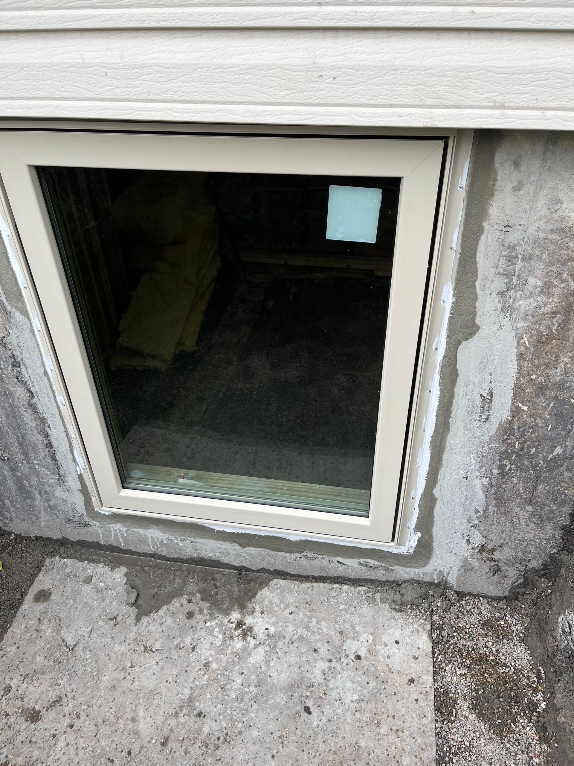 A tan-framed basement window installed in a concrete foundation, showing an interior view of a dark space.