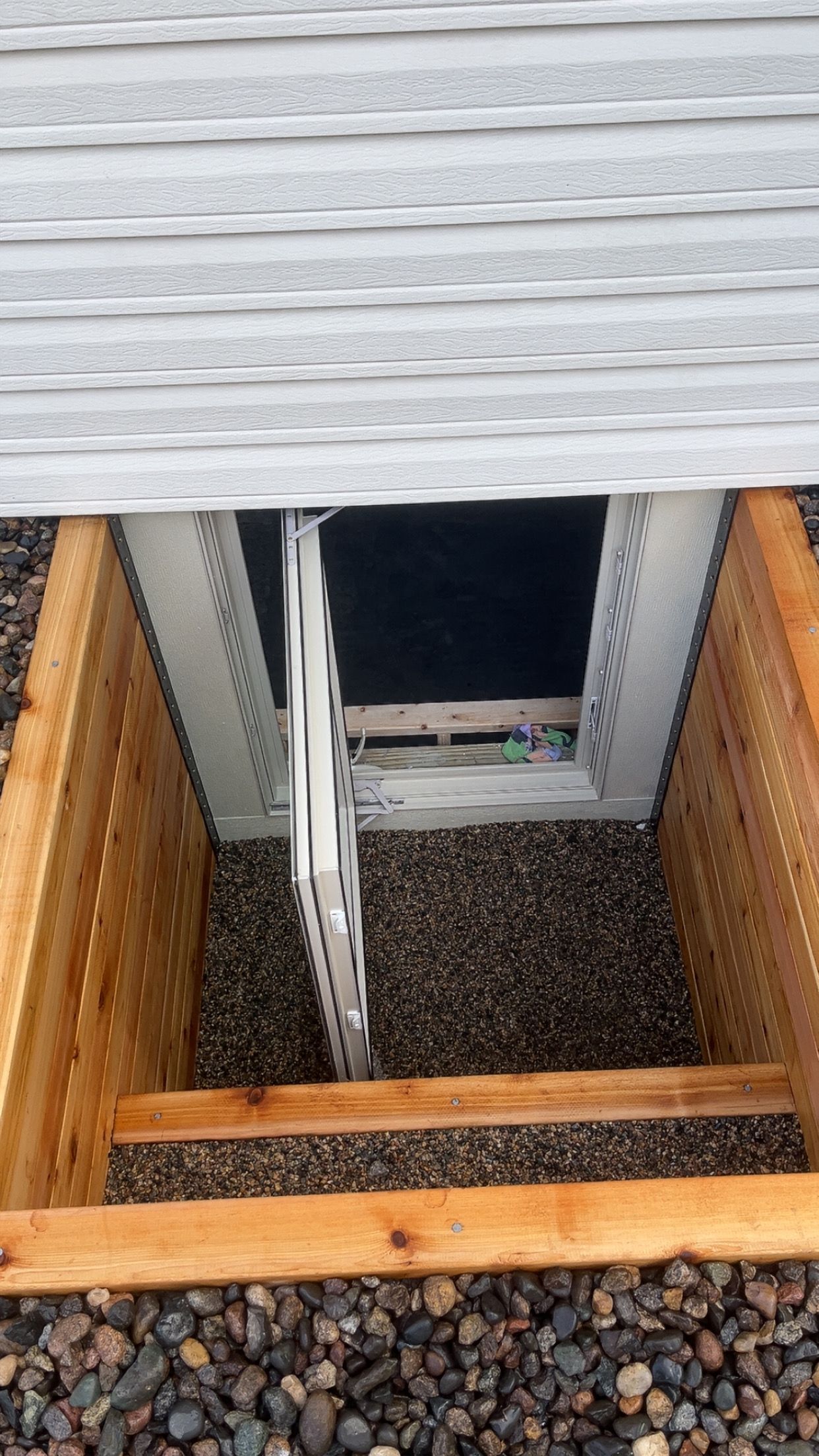 An egress window well featuring a white emergency escape ladder, wooden walls, and a gravel-filled floor beneath siding.