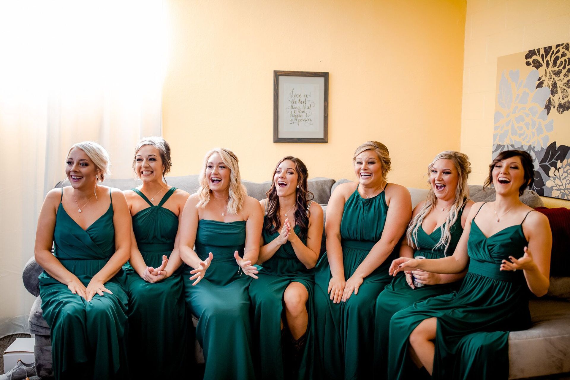Bridesmaids in emerald green dresses, excited and laughing, seated on a couch.